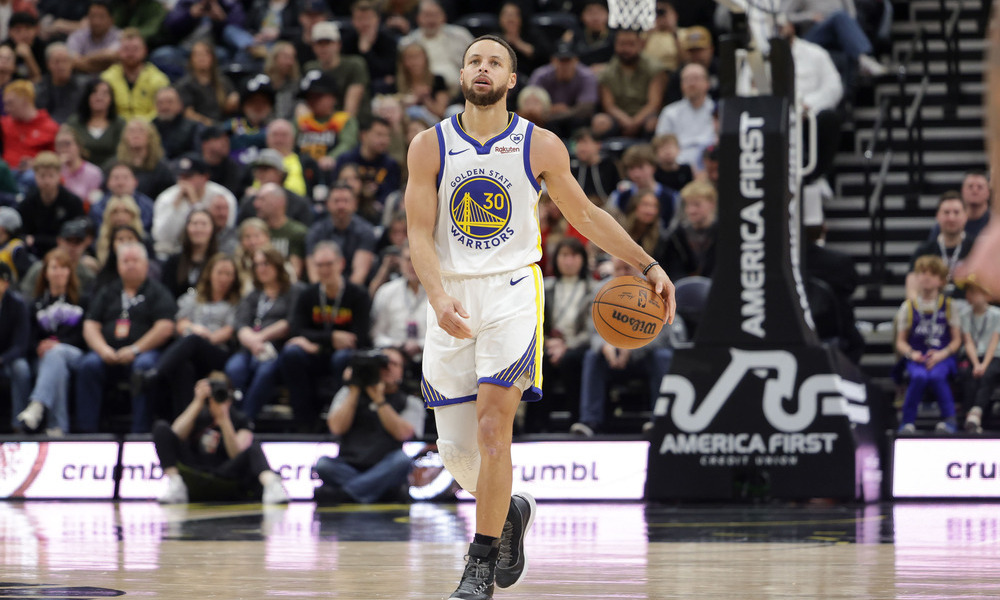 Steph Curry dribbling the ball up the floor as he looks up to the scoreboard.